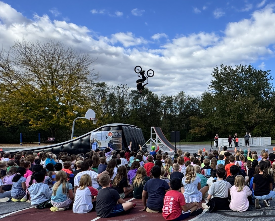 A large group of Monroe Elementary students sit outside on the playground, watching a BMX stunt show. In the background, a BMX rider is performing a high jump in midair against a bright blue sky with scattered clouds. The setup includes ramps and safety mats, and students are looking up in excitement. Trees with early autumn leaves frame the scene.