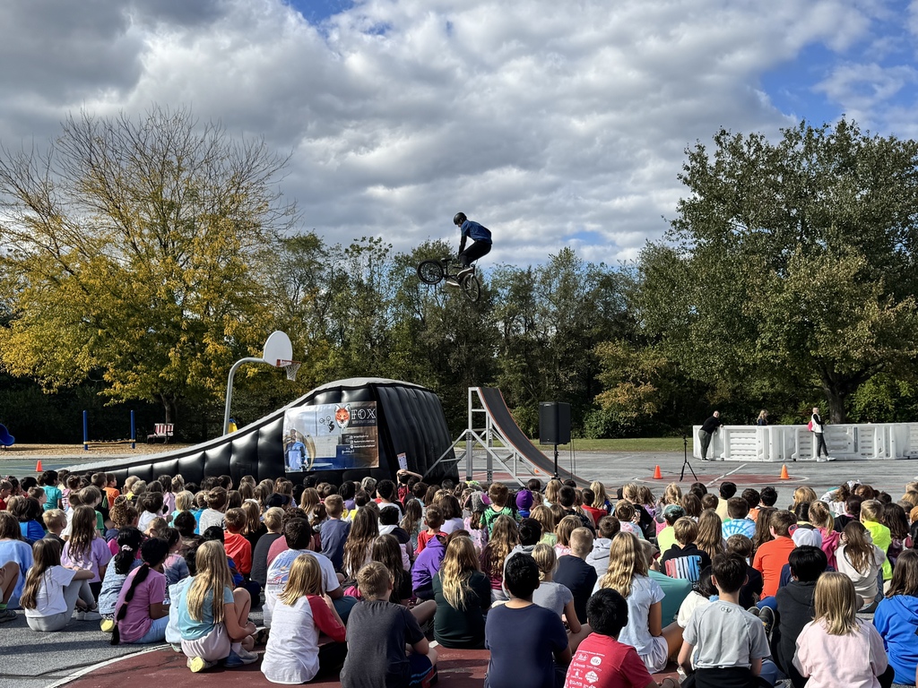 Monroe Elementary students sit outdoors watching a BMX stunt show. A BMX rider soars high in the air above a ramp while students look on in excitement. The show takes place under a partly cloudy sky with trees showing early fall colors in the background.