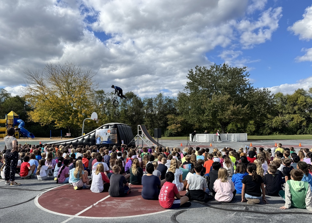 Monroe Elementary students sit outdoors watching a BMX stunt show. A BMX rider soars high in the air above a ramp while students look on in excitement. The show takes place under a partly cloudy sky with trees showing early fall colors in the background.