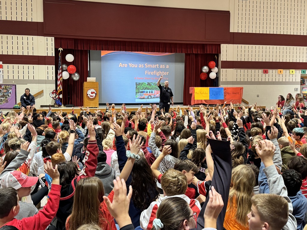 Monroe Fire Department's Chief Swab speaks to a large group of elementary students at assembly in Multi-Purpose Room.  He is in front of stage, in background are balloons, podium, and table set up for "Are You as Smart as a Firefigher" game.