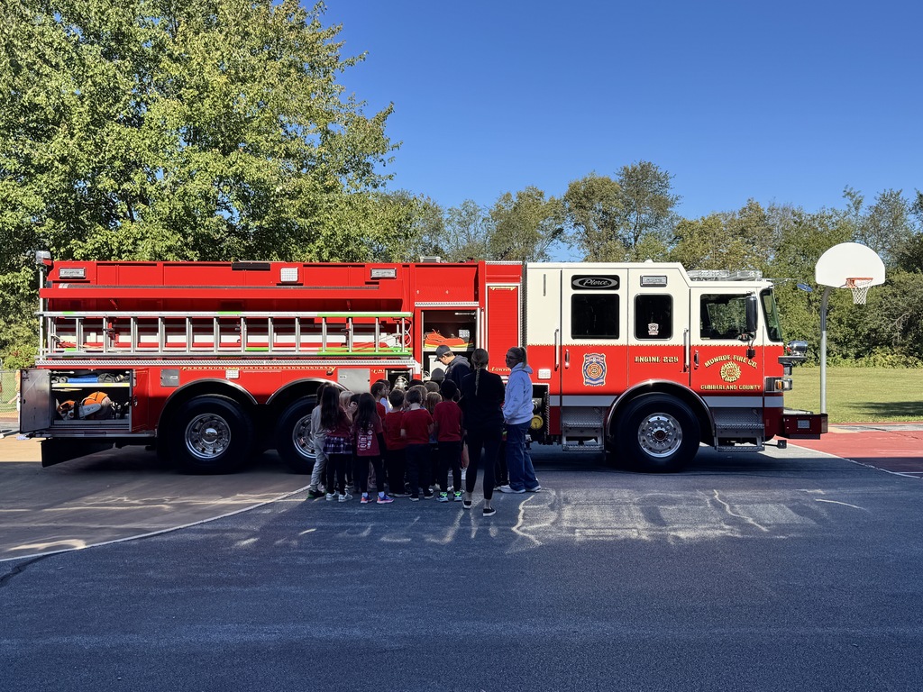 A group of elementary school students stands with a fireman with Monroe Fire Department fire truck parked near a tree and field on a bright sunny day.  Basketball hoop is visible in background.