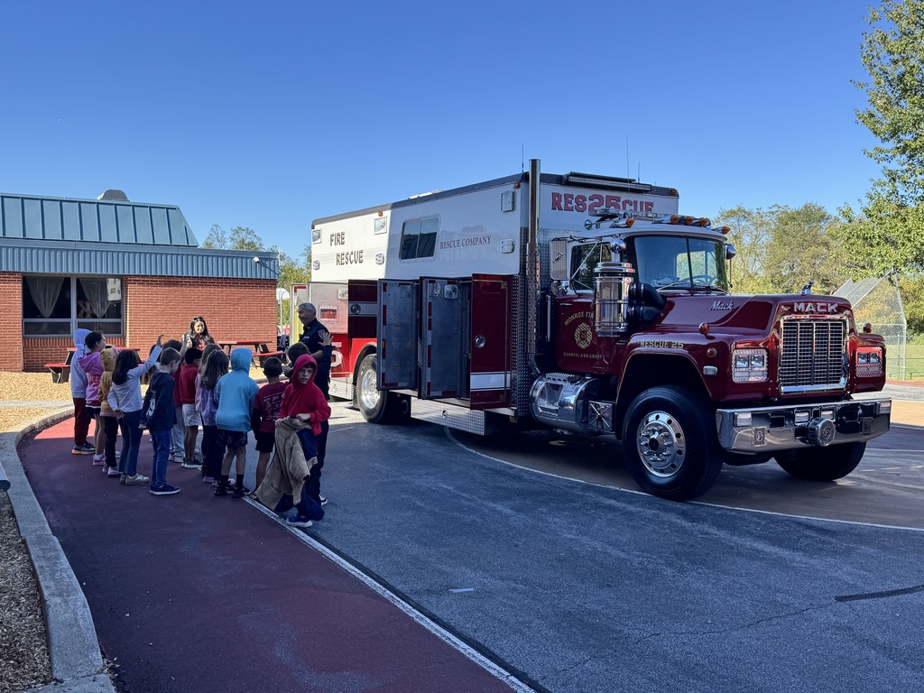 A group of elementary school students walks in a line past a Monroe Fire Department rescue truck parked outside a brick school building. A firefighter stands nearby talking with the children on a sunny day. Red picnic tables are visible in the background.