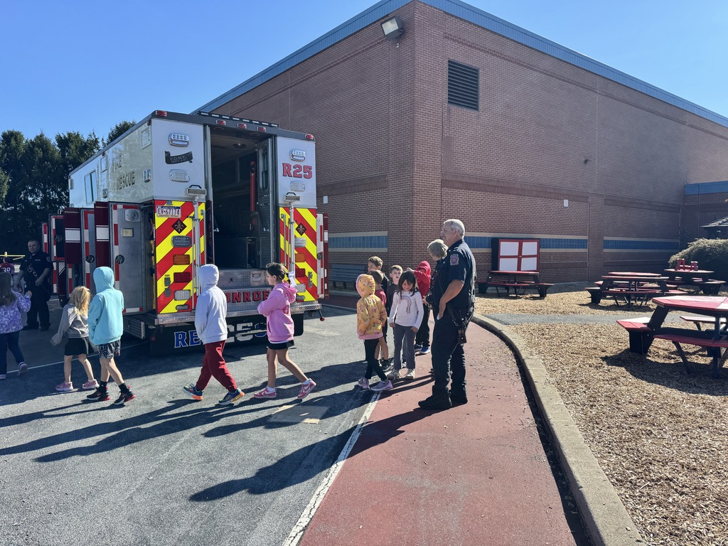 A group of elementary school students walks in a line past a Monroe Fire Department rescue truck parked outside a brick school building. A firefighter stands nearby talking with the children on a sunny day. Red picnic tables are visible in the background.