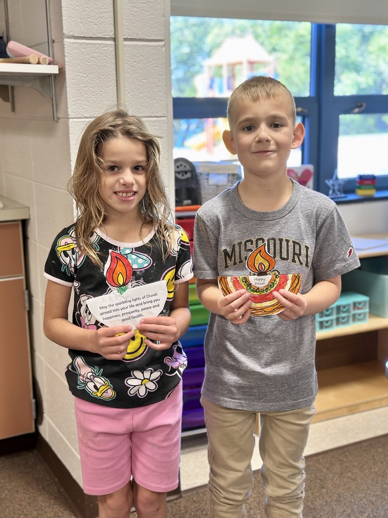 Two children in a classroom holding colorful Diwali crafts.