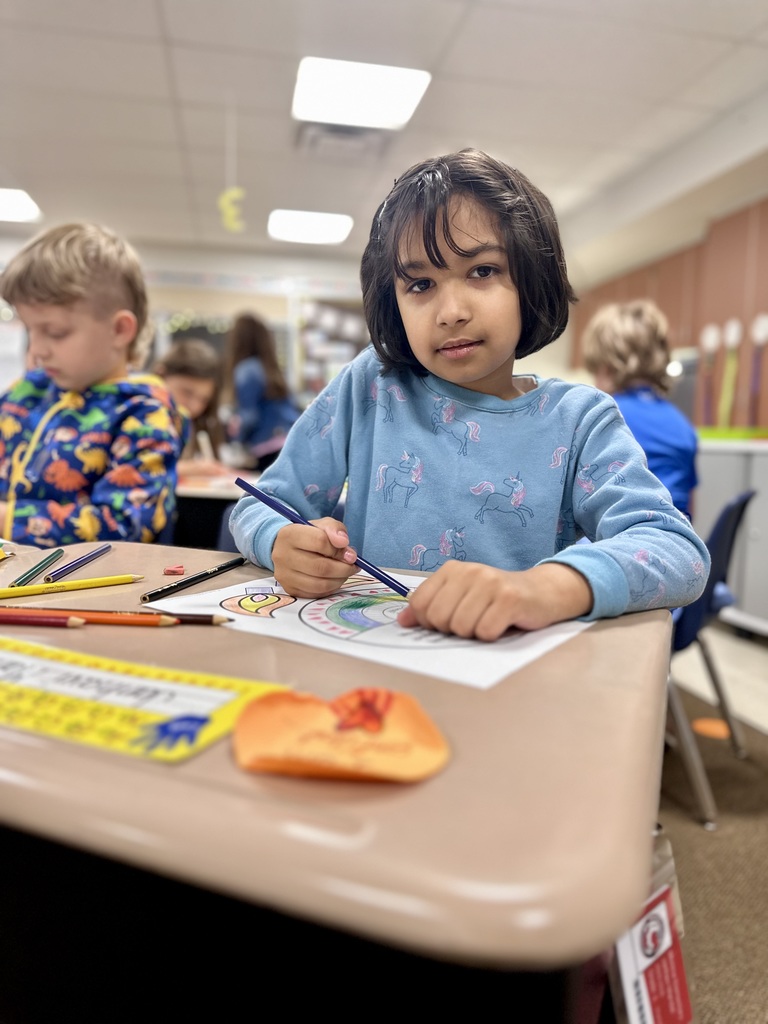 Child in a classroom coloring at a desk with colored pencils, wearing a blue unicorn shirt; other children are working in the background.