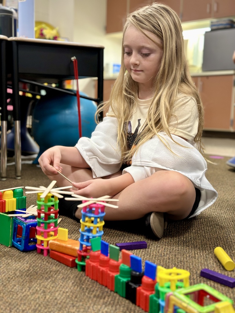 Child sitting on a classroom floor, building a colorful Great Wall of China with plastic blocks and wooden sticks.
