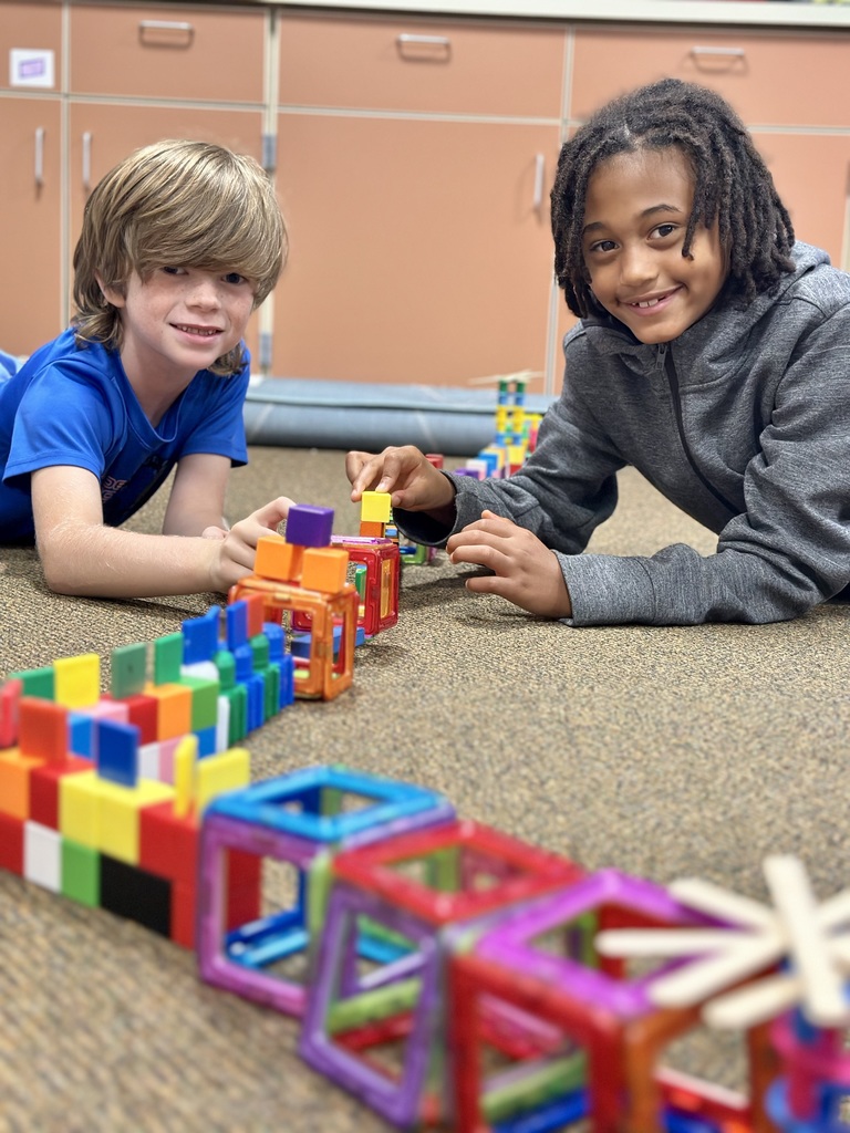 Two children are lying on a classroom floor, smiling and playing with colorful building blocks and magnetic tiles to build their version of the Great Wall of China.