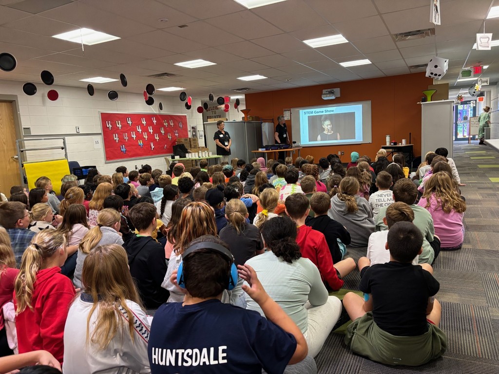 Children seated on the floor in a classroom, watching a presentation titled "STEM Club Show" with two presenters standing near a screen and equipment.