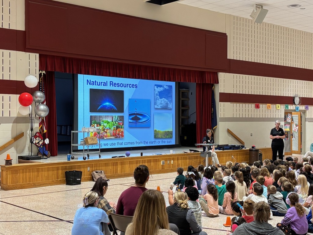 Children gathered in a classroom, viewing a presentation on "Natural Resources" with visuals of Earth, water, clouds, produce, and fields; two presenters are at the front.