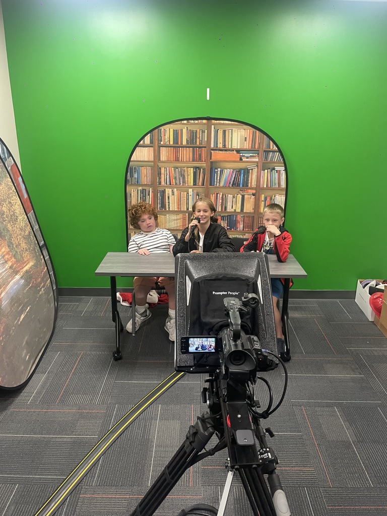 Three elementary students sit at a table in front of a green screen with a bookshelf prop, as part of a filming setup for Middlesex Elementary's LionTV.