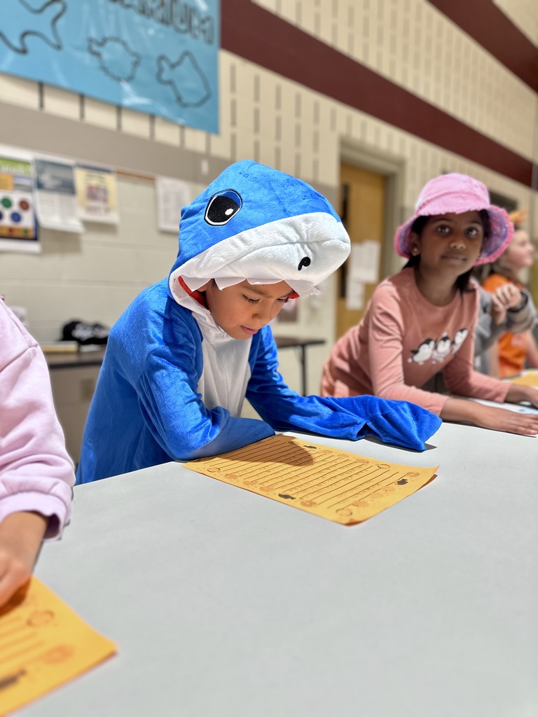 A student in a blue shark costume reads from an orange paper at a table next to another child in a pink outfit, with a fish-themed banner in the background.