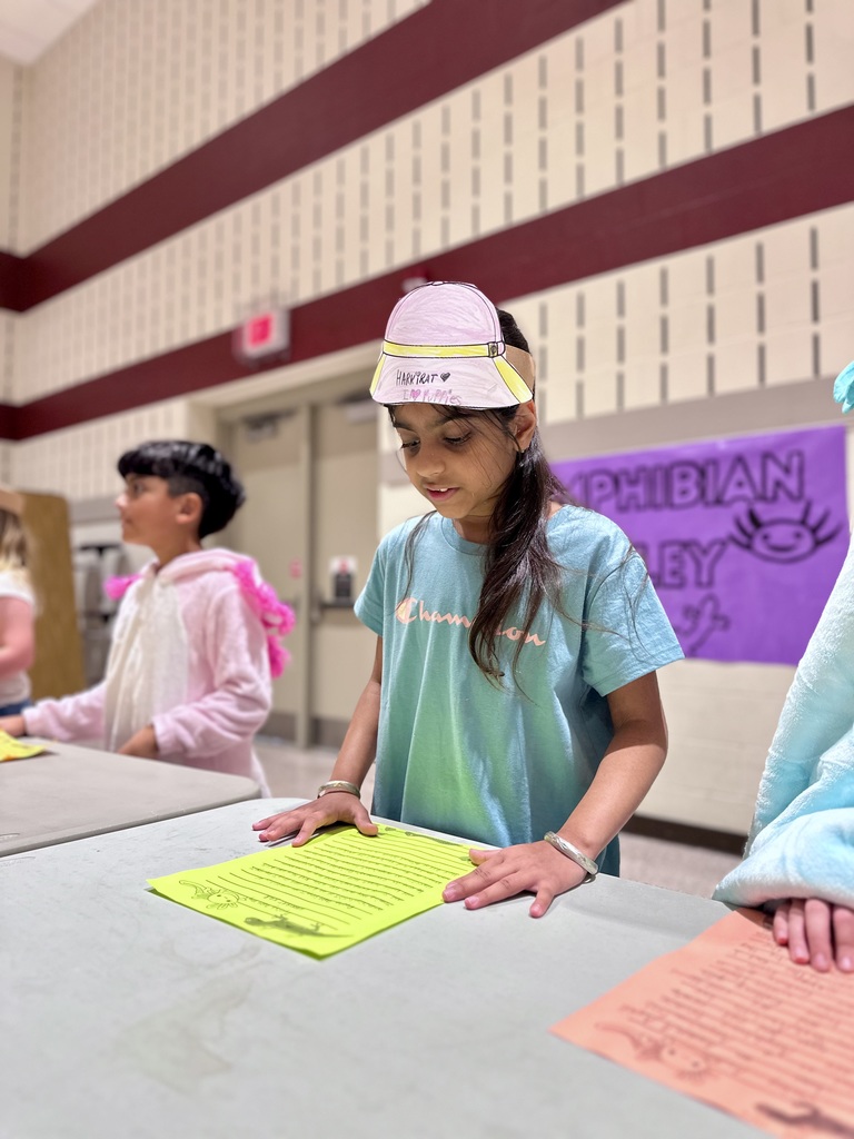 A student wearing a "Panther" visor reads from a yellow paper at a table near a purple "AMPHIBIAN ALLEY" poster.