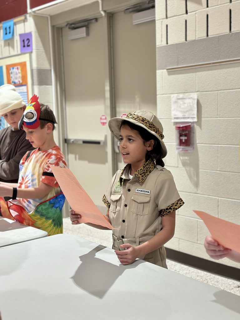 Children stand at a table holding orange papers; one child in front wears a safari outfit with a "Zookeeper" name tag.
