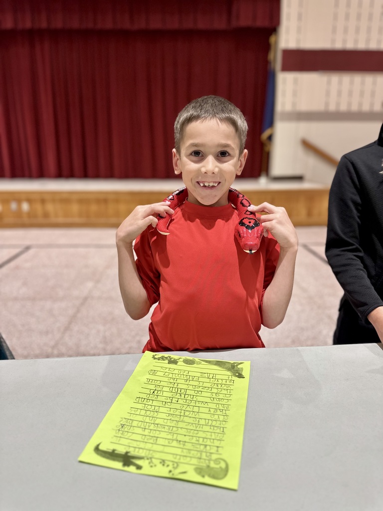 A student in a red shirt holds a red and black plush snake around their neck while standing behind a table with a yellow handwritten sign.