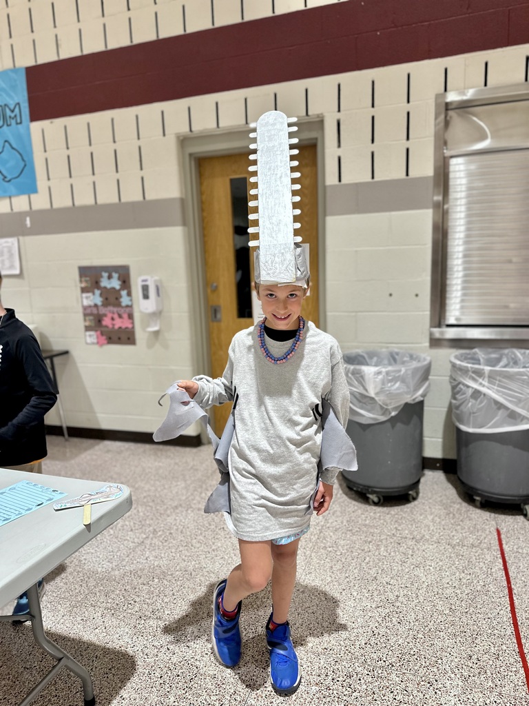 A student wearing a sawfish costume with a tall headpiece and fin-like attachments stands in a school cafeteria.