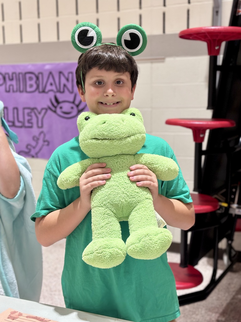 A student in a green shirt and frog headband holds a frog plush toy in front of a banner that reads "AMPHIBIAN ALLEY."
