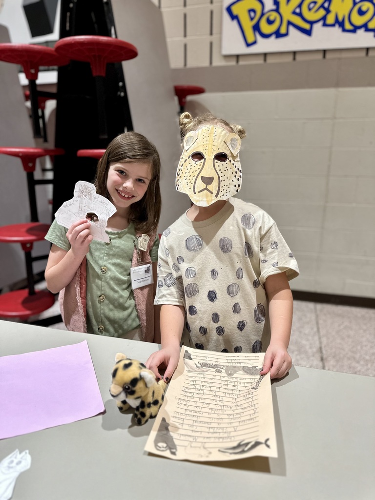 Two students in front of table, one wearing a mask and the other showing her mask.