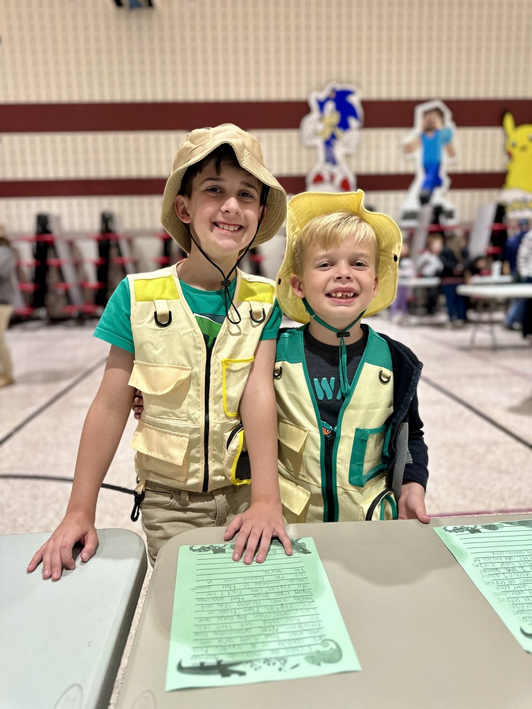 Two students with zoo keeper costumes on, smiling in front of their table.