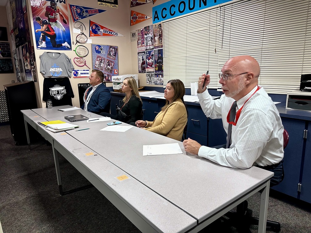 Four individuals sit at a table with papers and a laptop, acting as the Sharks for a classroom Shark Tank project.