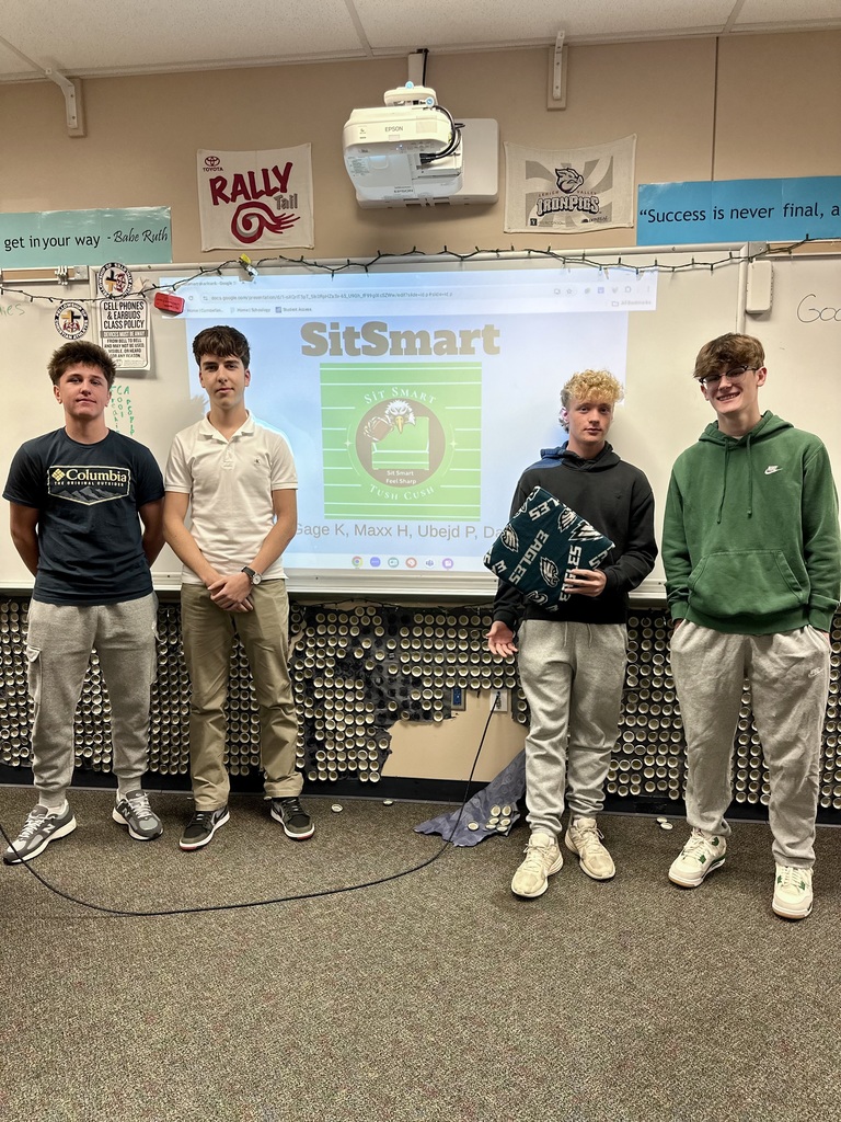 Four students stand in front of a classroom whiteboard presenting their Shark Tank idea, "SitSmart," with a slide showing the logo and their names.