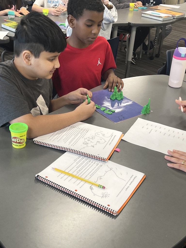 Students working at a classroom table with Play-Doh and notebooks, shaping green pieces on a purple mat while following an illustrated activity guide.