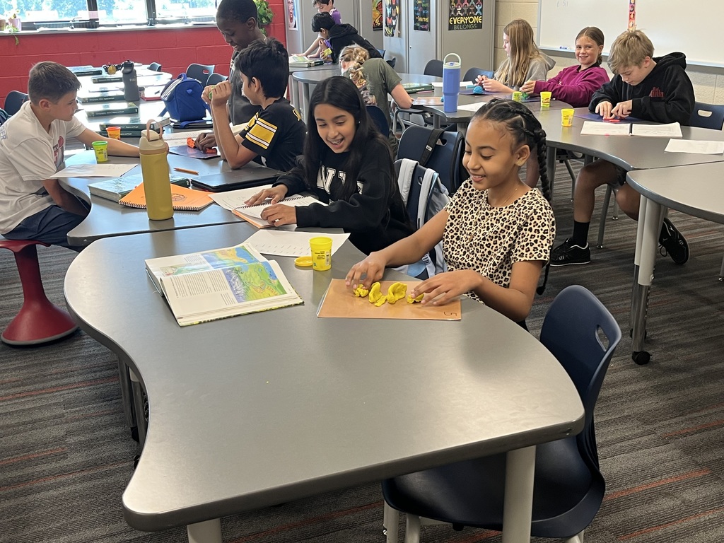 Students seated at curved tables in a classroom, working on activities with notebooks and yellow modeling clay. A book and water bottle are visible on the front table, and colorful posters decorate the walls in the background.
