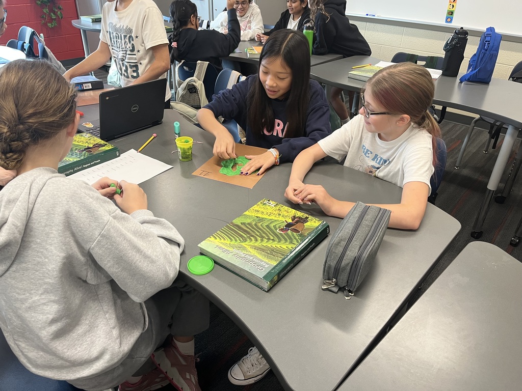 Students sitting at a classroom table working with green modeling clay and textbooks. A pencil case, notebook, and laptop are also visible on the table. Other students and desks are in the background.