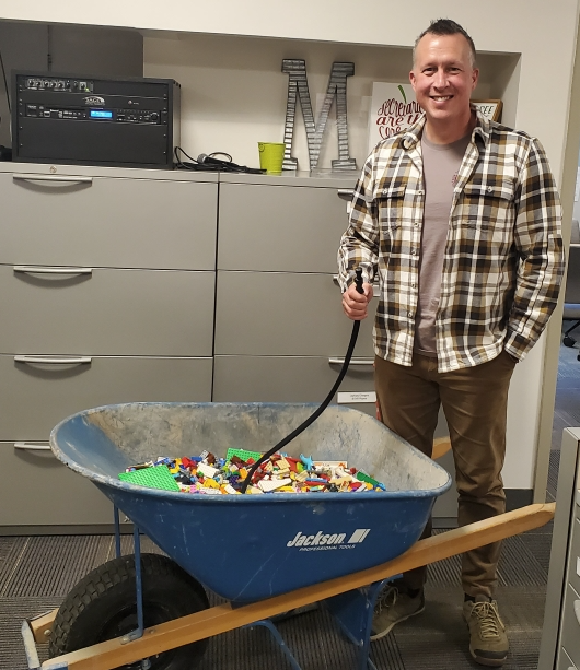 Male teacher drinking from a hose that is housed in a Lego-filled wheelbarrow.