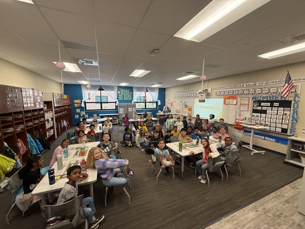 Large group of Winding Creek Elementary students in the Pages & Pals group gathered in a bright classroom, sitting at tables and on the floor, with colorful decorations, cubbies, and learning materials visible.