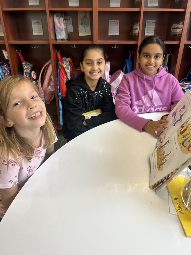 Three Winding Creek Elementary students from the Pages & Pals group sit at a round table reading a colorful picture book, with cubbies and backpacks in the background.