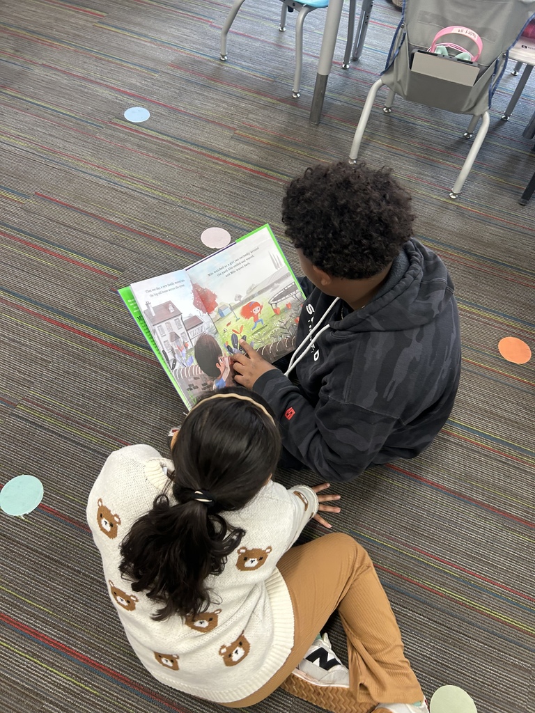 Two Winding Creek Elementary students from the Pages & Pals group sit on a classroom carpet reading a colorful picture book together.