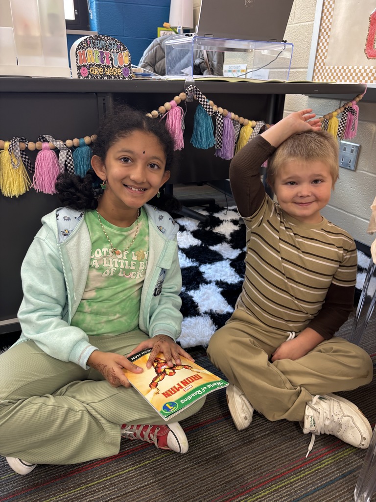 Two Winding Creek Elementary students from the Pages & Pals group sit on the floor in a cozy reading nook, with one holding a book