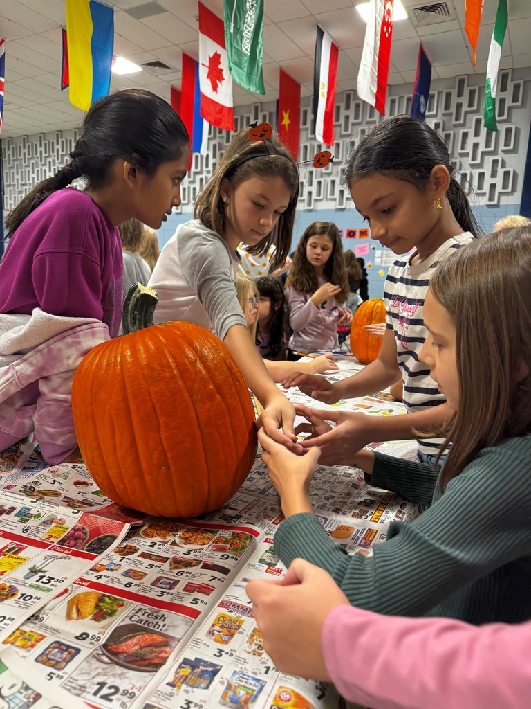Several Hampden Elementary 4th graders gather around a table covered with newspaper ads, examining and handling pumpkin seeds and pulp during a pumpkin science activity. A large orange pumpkin sits prominently on the table, and colorful international flags hang from the ceiling in the background.