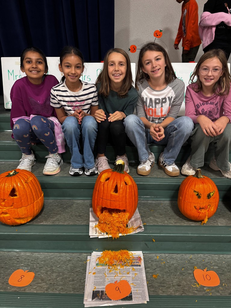Five Hampden Elementary 4th graders sit on green steps behind three carved pumpkins. The pumpkins have creative faces, with the middle one designed to look like it is vomiting pumpkin seeds and pulp onto newspaper. Orange paper cutouts with numbers are placed in front of each pumpkin.