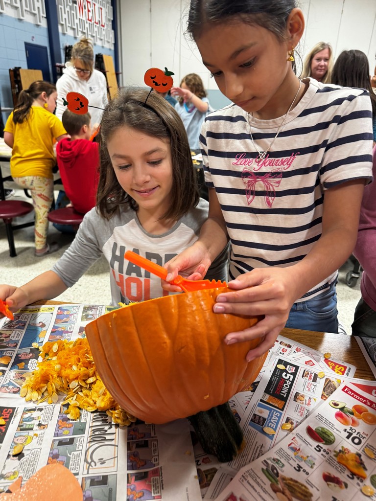 Two Hampden Elementary 4th graders scoop seeds and pulp from a large orange pumpkin using orange plastic tools. The pumpkin is resting on a table covered with newspaper ads, and piles of seeds and stringy pulp are visible next to it. Other students and adults are seated at tables in the background, participating in similar pumpkin science activities.