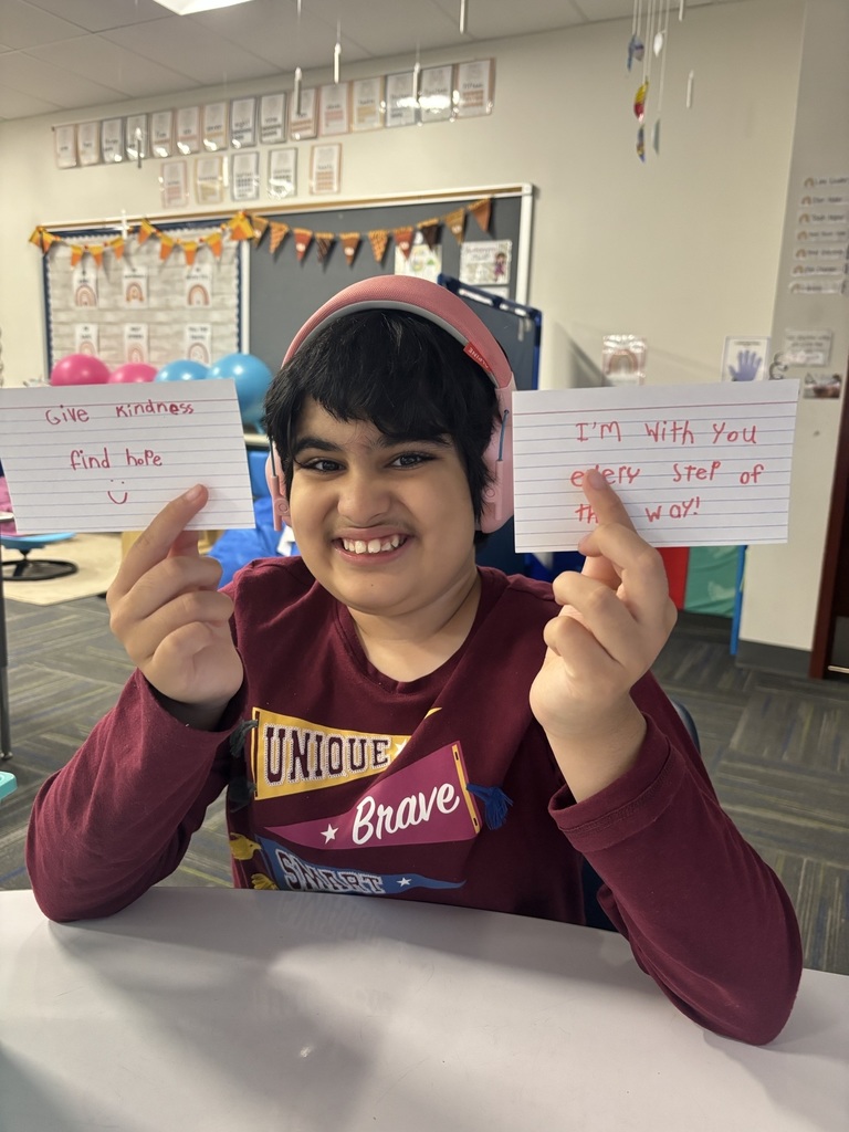 Student holding two handwritten kindness notes in a classroom; one reads “Give kindness, find love :)” and the other says “I’m with you every step of the way.”