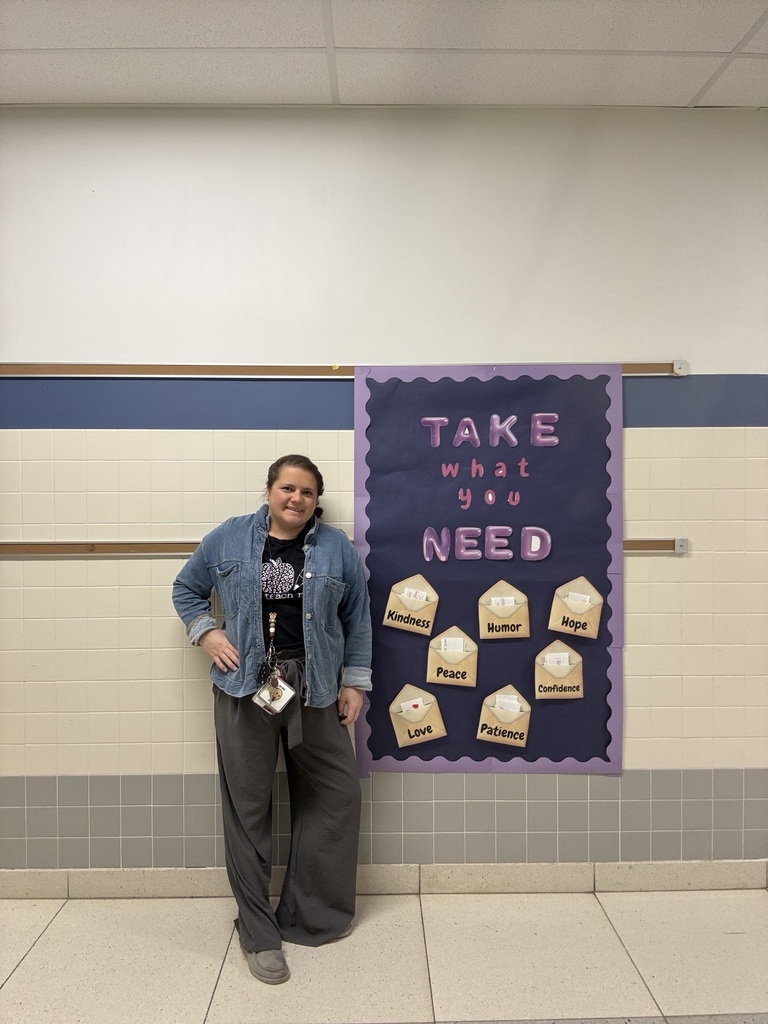 Teacher standing next to a purple bulletin board titled “Take what you need,” featuring envelopes labeled with words like Love, Peace, Patience, Kindness, Humor, and Hope.