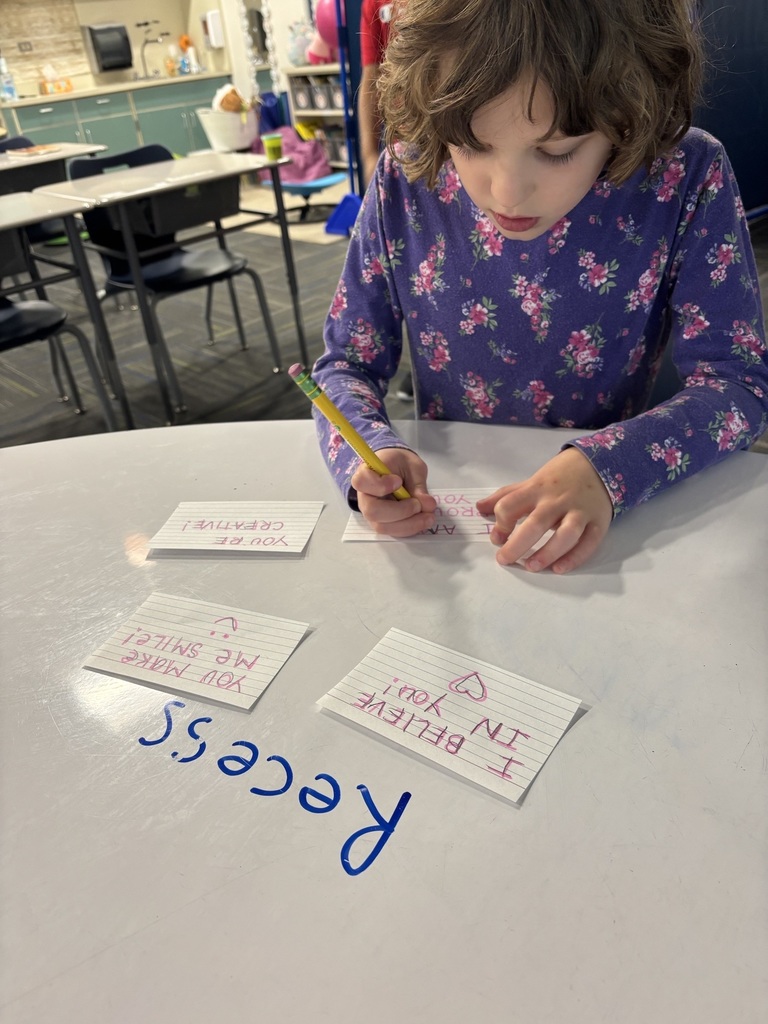 Student writing a kindness note at a table with other handwritten notes that read “I believe in you,” “You share the sparkle,” and “You are creative.”
