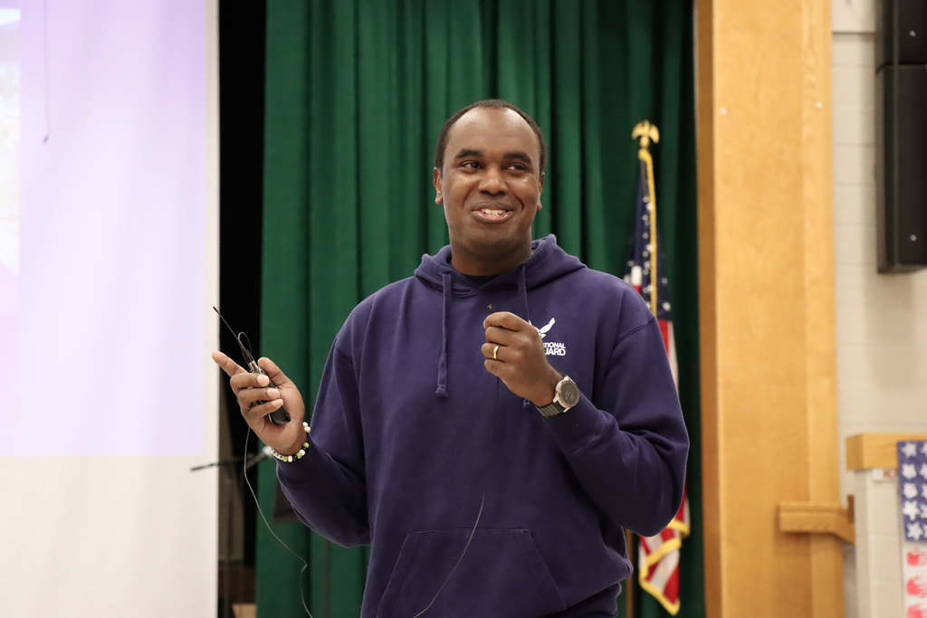 Veteran speaking during a Veterans Day event, holding a microphone and standing in front of a green curtain with an American flag visible in the background.