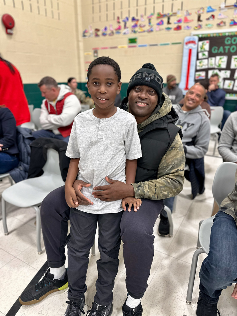 Adult seated in a chair with a child standing in front, surrounded by other attendees in a school setting during a Veterans Day assembly.