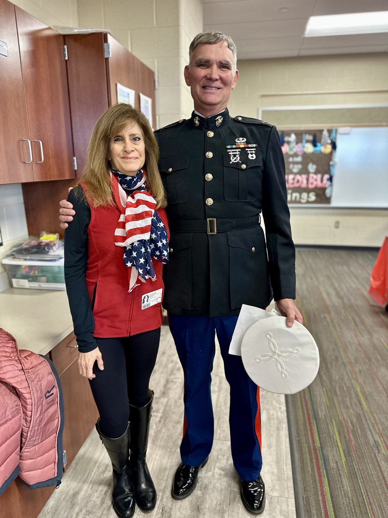 Person in a formal military dress uniform stands next to another person wearing a red vest and an American flag scarf inside a classroom setting. The person in uniform is holding a white military hat.