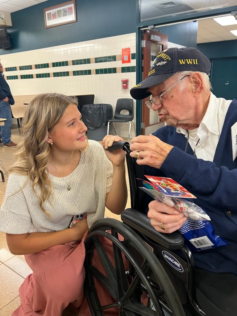 A person seated in a wheelchair wearing a WWII veteran cap holds a small gift bag while another person kneels beside them, holding the wheelchair armrest. They are indoors in a room with chairs and tables in the background.