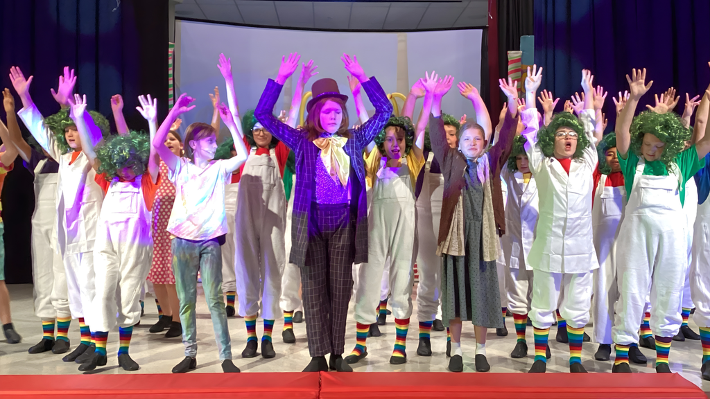 Children in colorful costumes performing on stage in a production of Willy Wonka Kids. The group includes a character dressed as Willy Wonka in a purple coat and top hat, surrounded by performers in white overalls, rainbow-striped socks, and green wigs, resembling Oompa-Loompas. All performers have their arms raised in unison under bright stage lights.