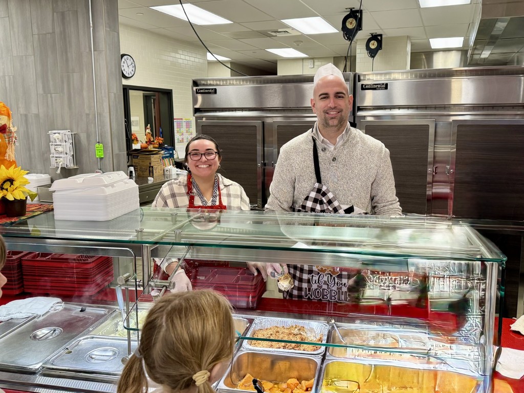 School administrators and district office staff assist food service crews in a cafeteria, serving Thanksgiving meals to students from behind a counter with trays of food.