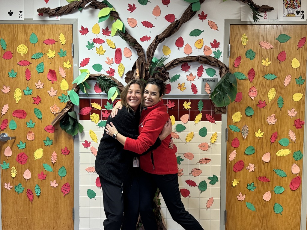Two teachers at Monroe Elementary School hug in front of the gratitude tree they created.