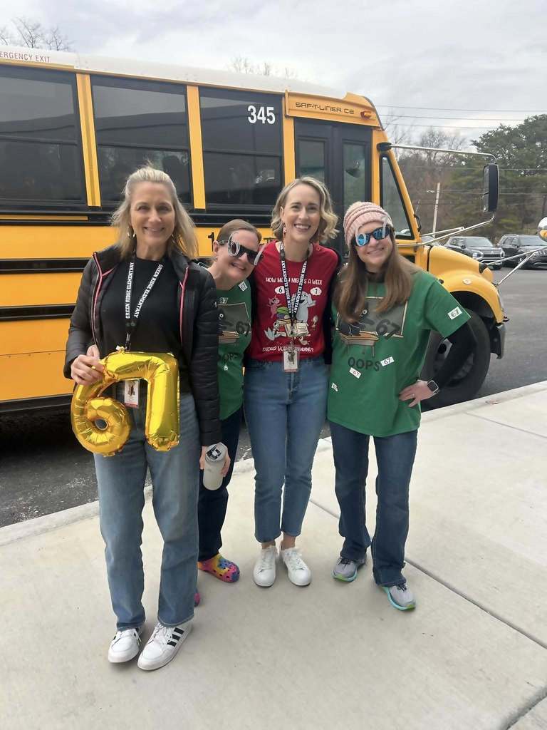 Four teachers standing in front of a yellow school bus, one holding gold balloon numbers “6” and “7,” celebrating the 67th day of school.