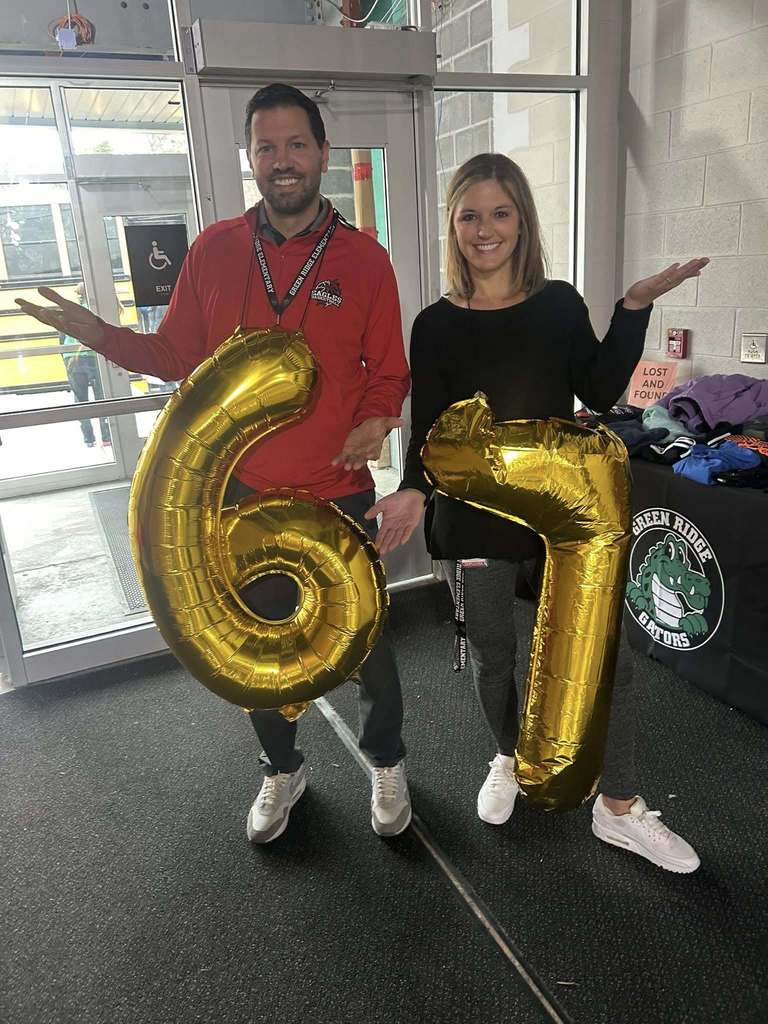 Green Ridge Admin holding large gold balloon numbers “6” and “7” to celebrate the 67th day of school, standing near an entrance with a Green Ridge Gators table in the background.