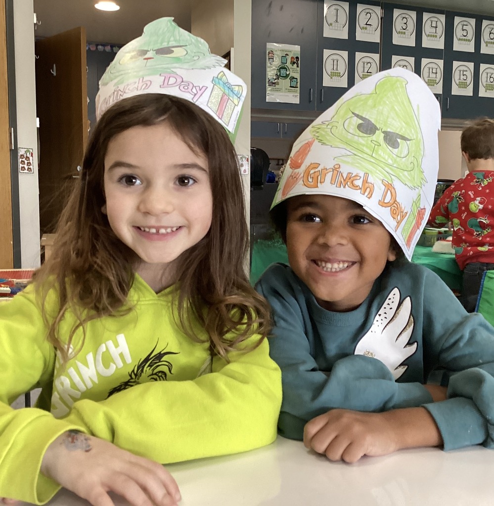 Two kindergarten students wearing Grinch-themed paper hats and festive outfits during Middlesex Elementary’s Grinch Day, seated at a classroom table.