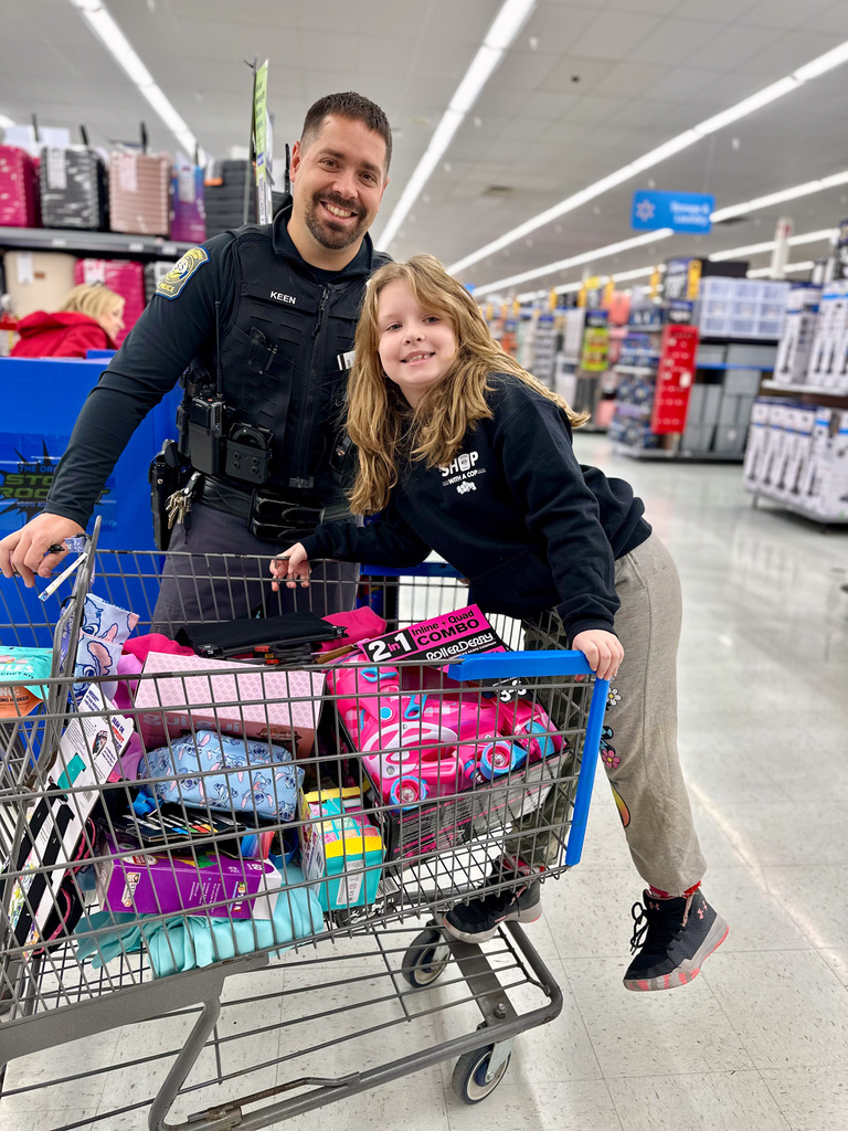 An officer and a child stand beside a shopping cart filled with clothing and toys during the 11th annual Shop with a Cop event.
