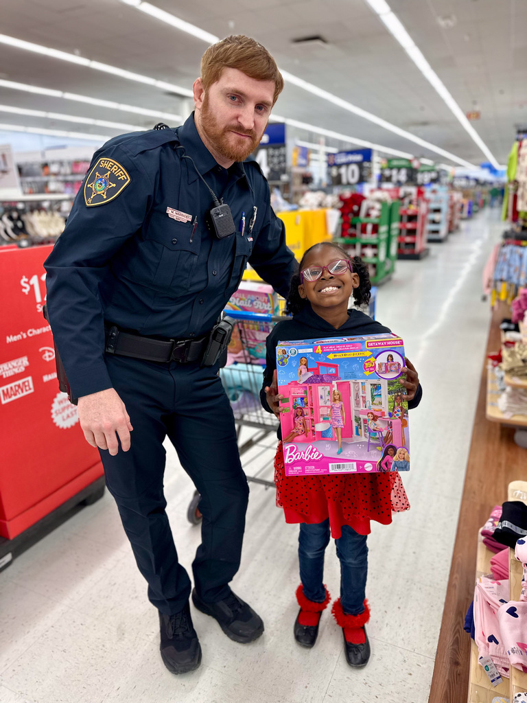 A sheriff’s deputy stands beside a child holding a Barbie playset during the 11th annual Shop with a Cop event.